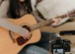 Woman sitting on a couch, filming themselves playing acoustic guitar.