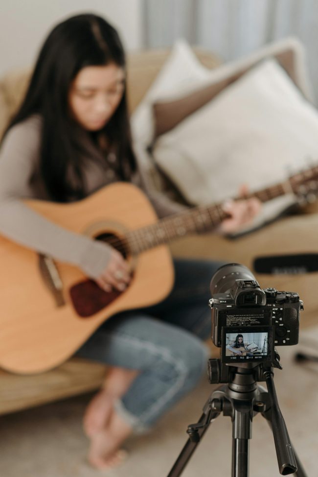 Woman sitting on a couch, filming themselves playing acoustic guitar.